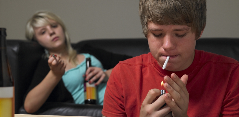 Young man smoking a cigarette with a girl in the background drinking a beer.