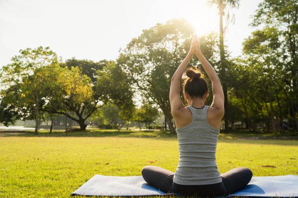 Woman sitting outside on a mat stretching her hands above her head.